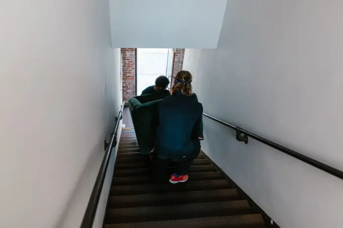 Två flyttare bär en grön fåtölj nerför en smal trappa mot en öppen ytterdörr